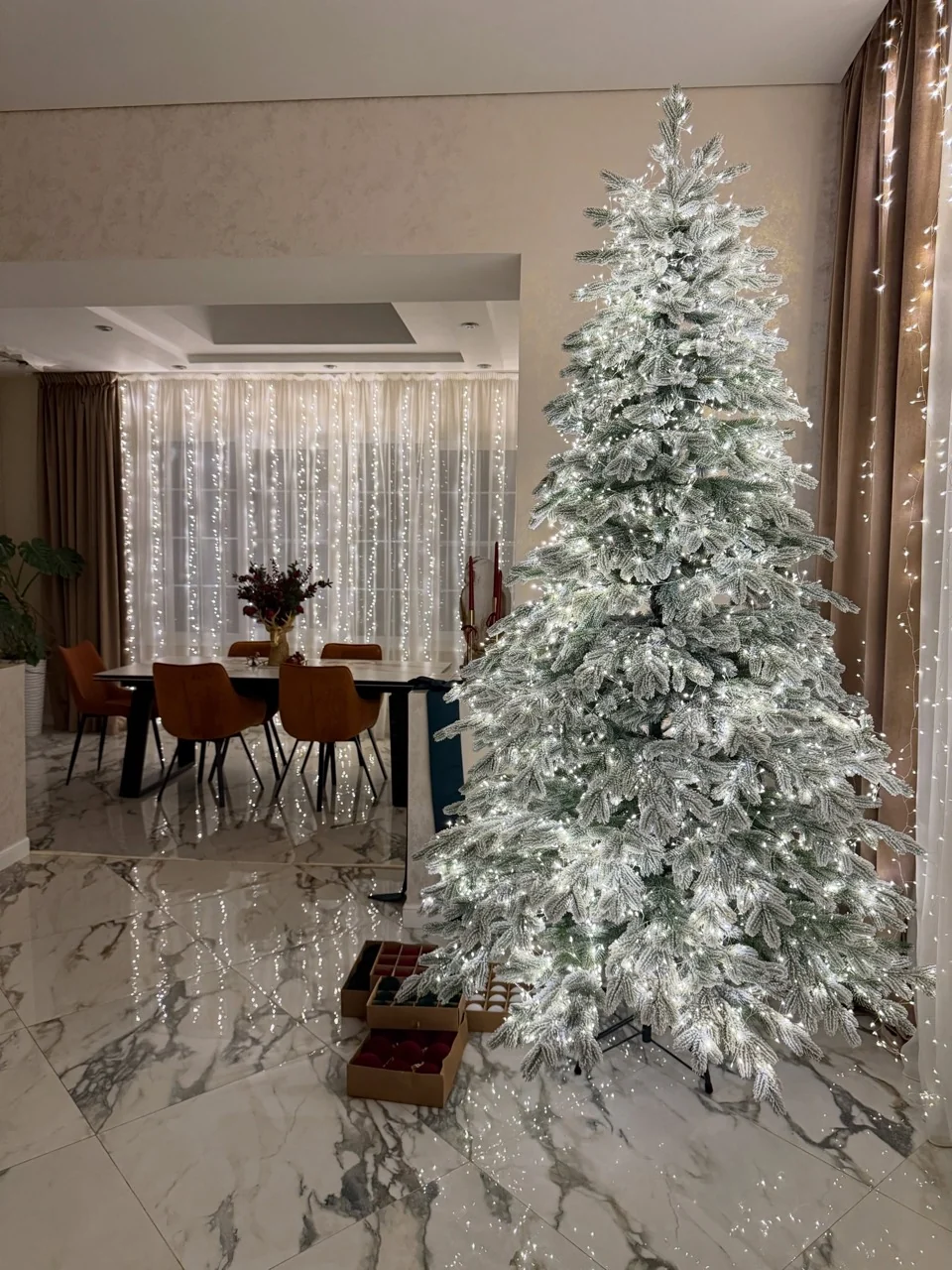 Illuminated flocked Christmas tree beside curtain lights, with ornament boxes on the marble floor.