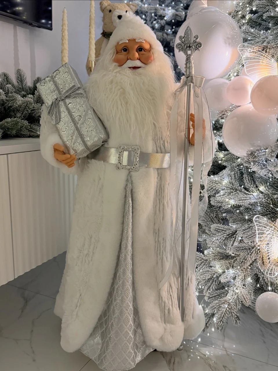 White-robed Santa decoration holding a silver present and staff beside a flocked Christmas tree.