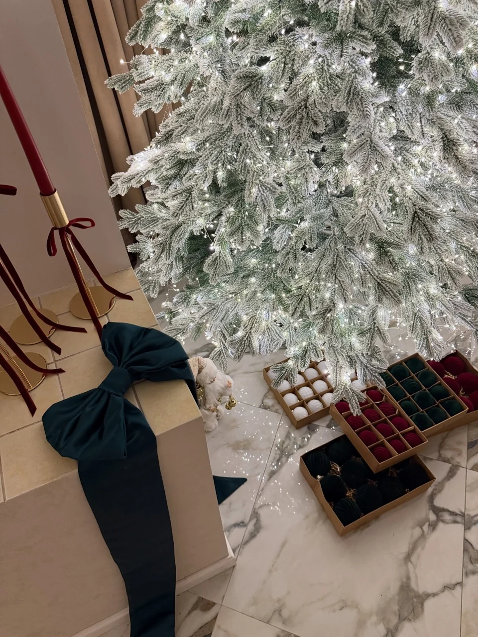 Flocked tree branches above boxes of burgundy, emerald and white ornaments beside a large emerald ribbon bow.