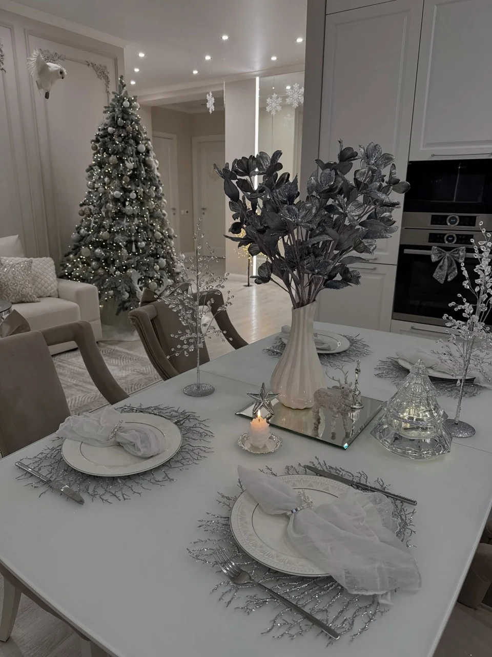 White and silver Christmas dining table with foliage and crystal decorations, with a decorated woodland tree beyond.