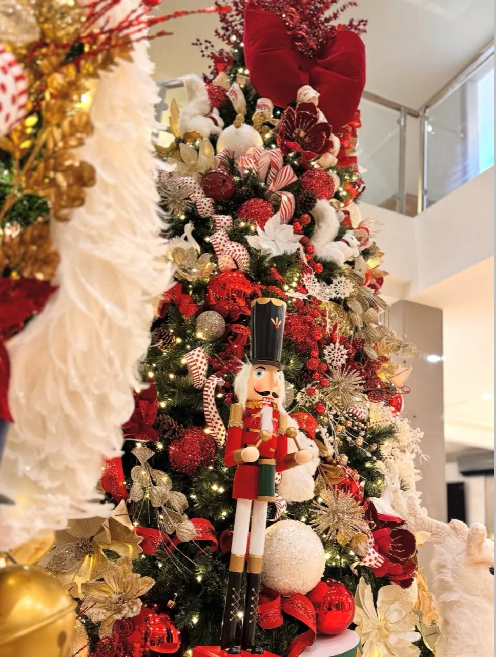 Red-uniformed nutcracker beside a Christmas tree filled with red baubles, gold accents, white ornaments and a large red bow.