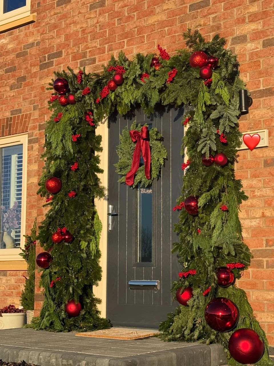 Front entrance surrounded by evergreen garlands, red ornaments and berries, with a red-bow wreath on a dark door.
