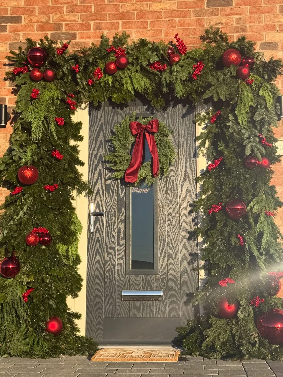 Evergreen Christmas garland framing a dark front door with red berries, large red baubles and a red velvet bow wreath.