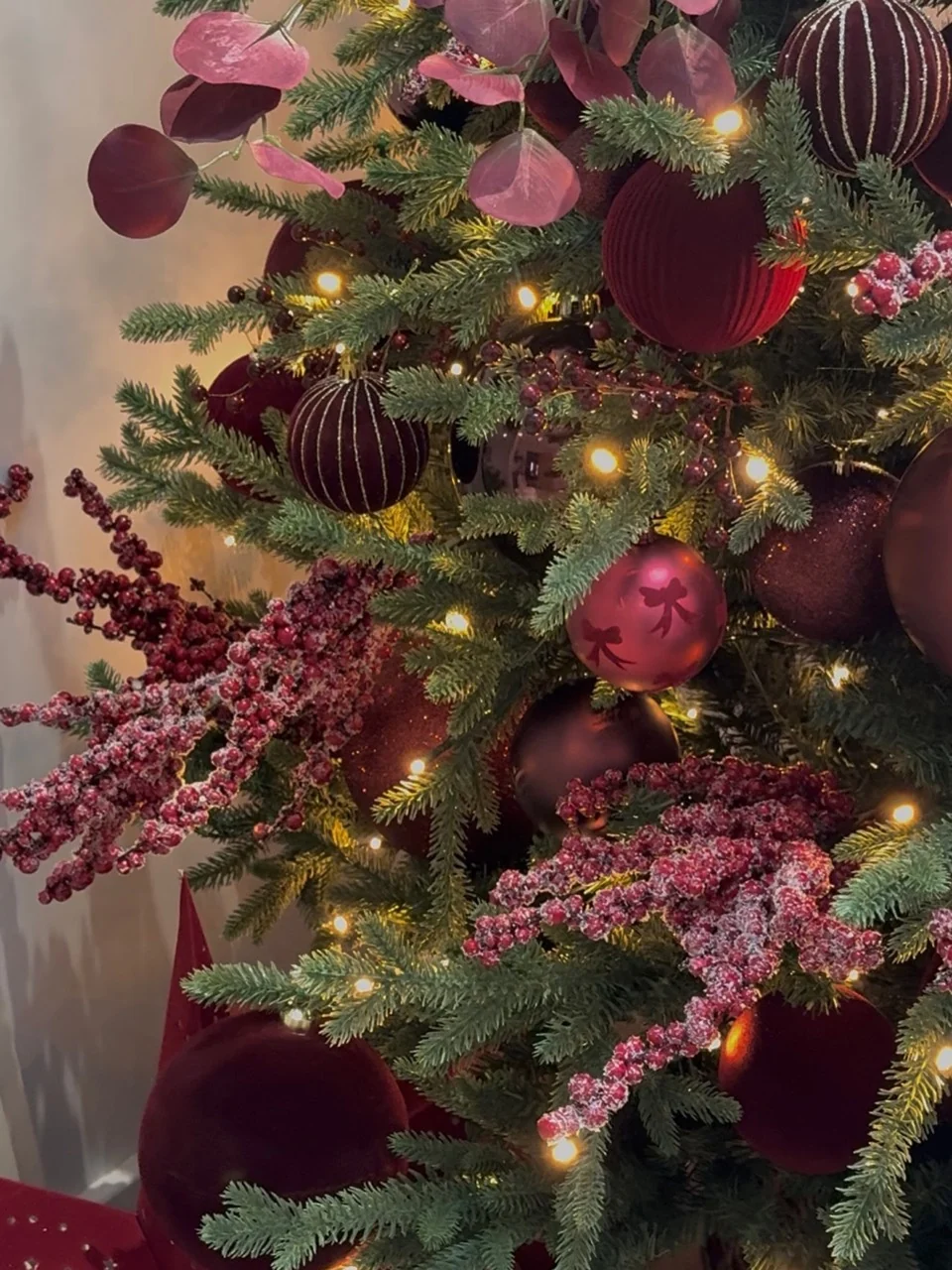 Close view of burgundy baubles, round purple leaves and frosted berry sprays among illuminated Christmas tree branches.
