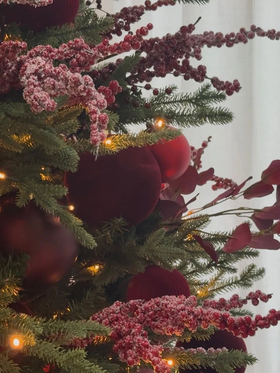 Velvet burgundy ornaments and frosted berry sprays woven through evergreen branches with warm fairy lights.