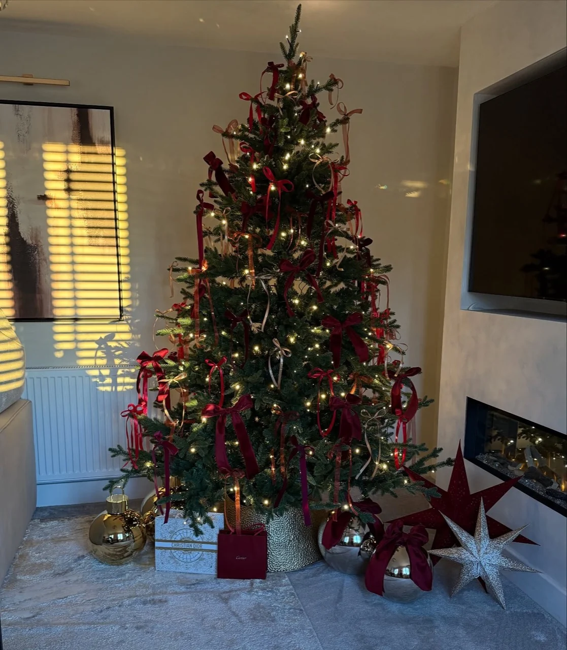 Christmas tree with burgundy and gold ribbon bows and warm lights beside a fireplace, with oversized metallic ornaments and stars at its base.