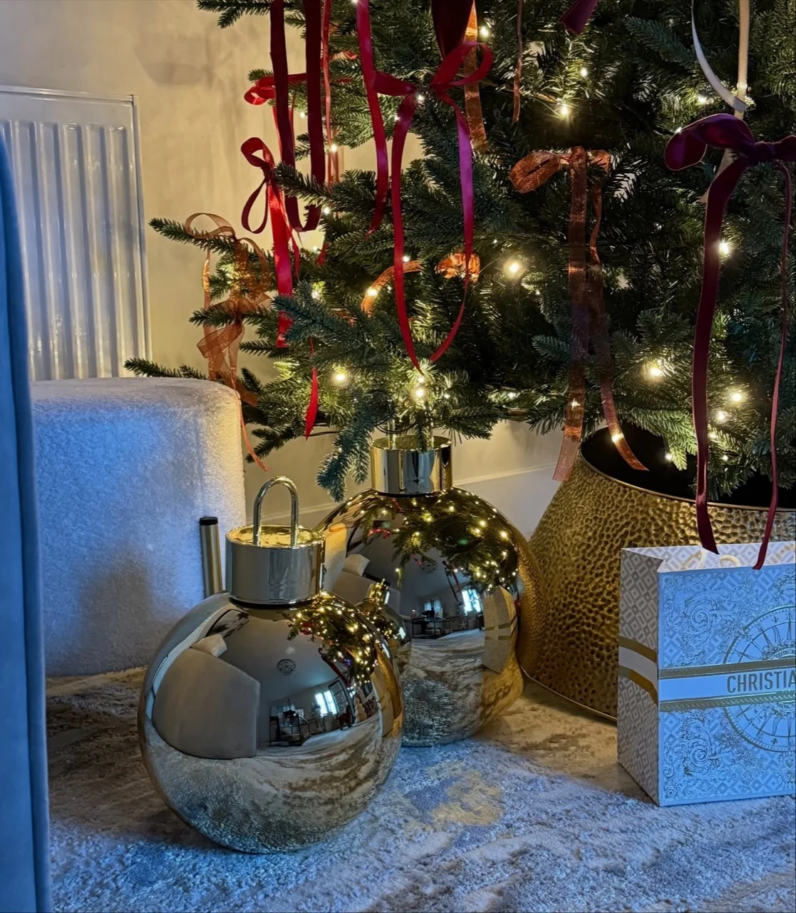 Oversized reflective metallic baubles beneath Christmas tree branches with burgundy and gold ribbon bows.