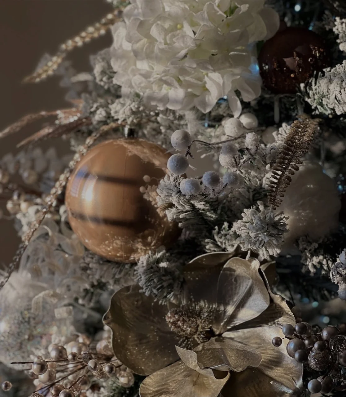 Gold Christmas bauble beside white flowers, pale berry sprays and frosted evergreen branches.