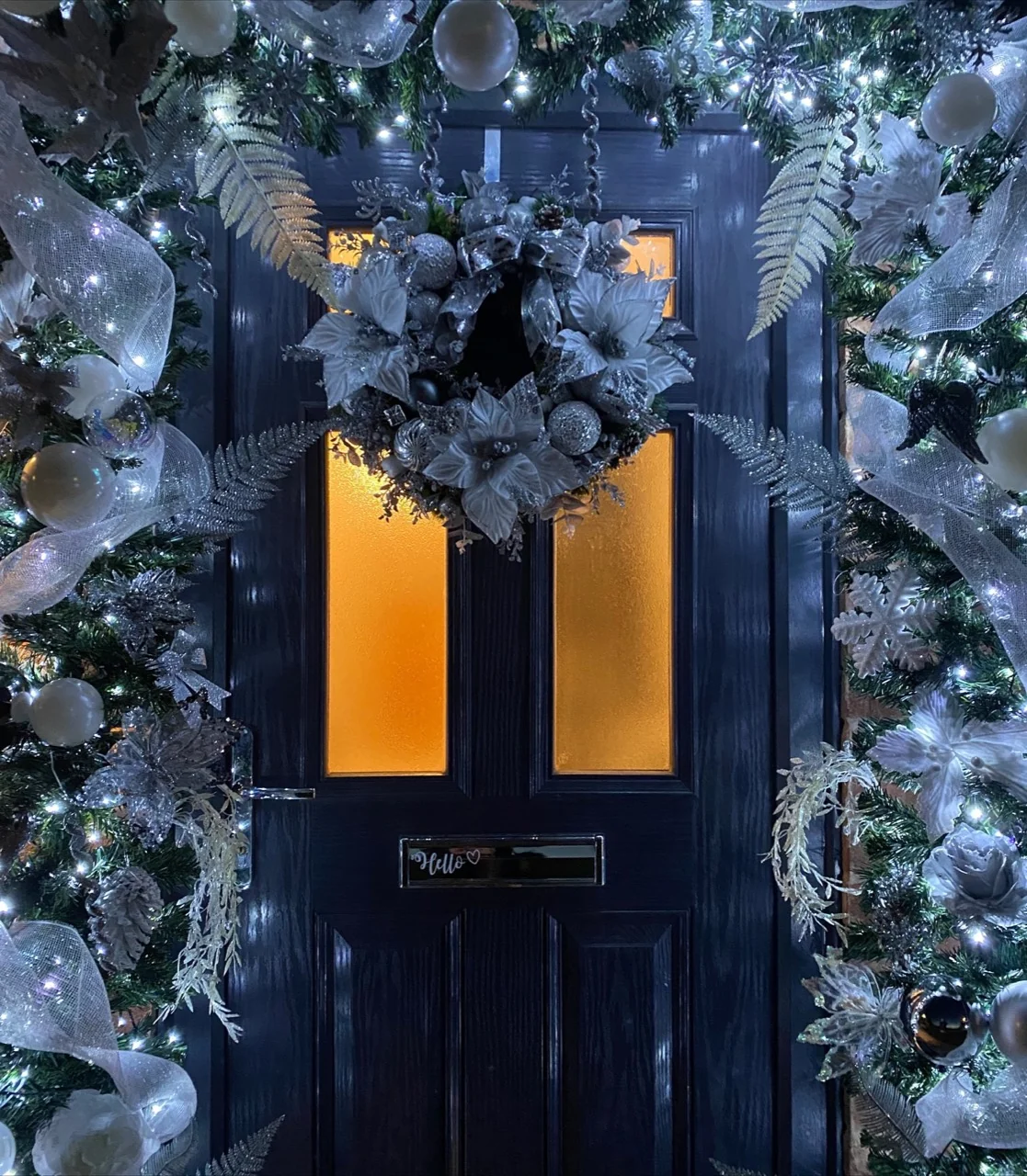 Dark front door framed by illuminated silver and white Christmas garlands with mesh ribbon, flowers, baubles and a matching wreath.